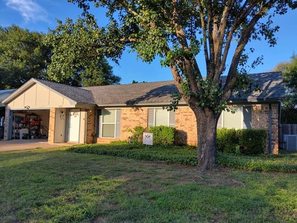 Brick house with a front yard and large tree, in a residential neighborhood. Bright sunlight.
