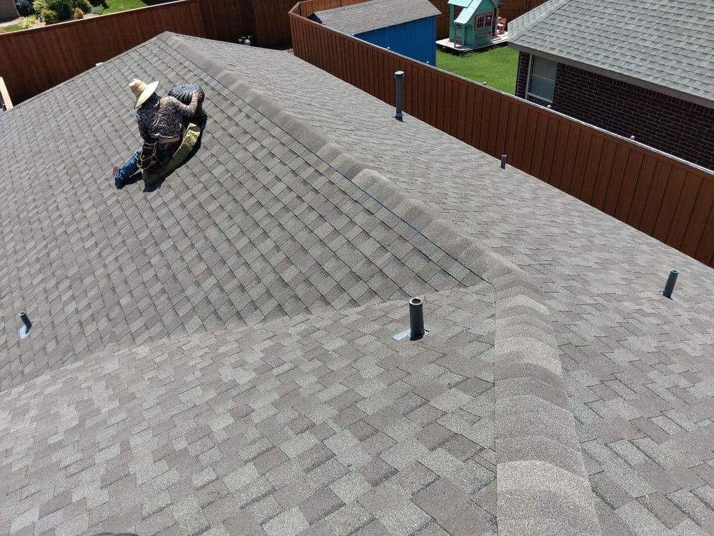 Roofer working on a sloped asphalt shingle roof with vents and neighboring houses in view.