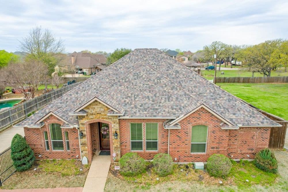 Aerial view of a brick home with a textured roof and landscaped yard in a suburban area.