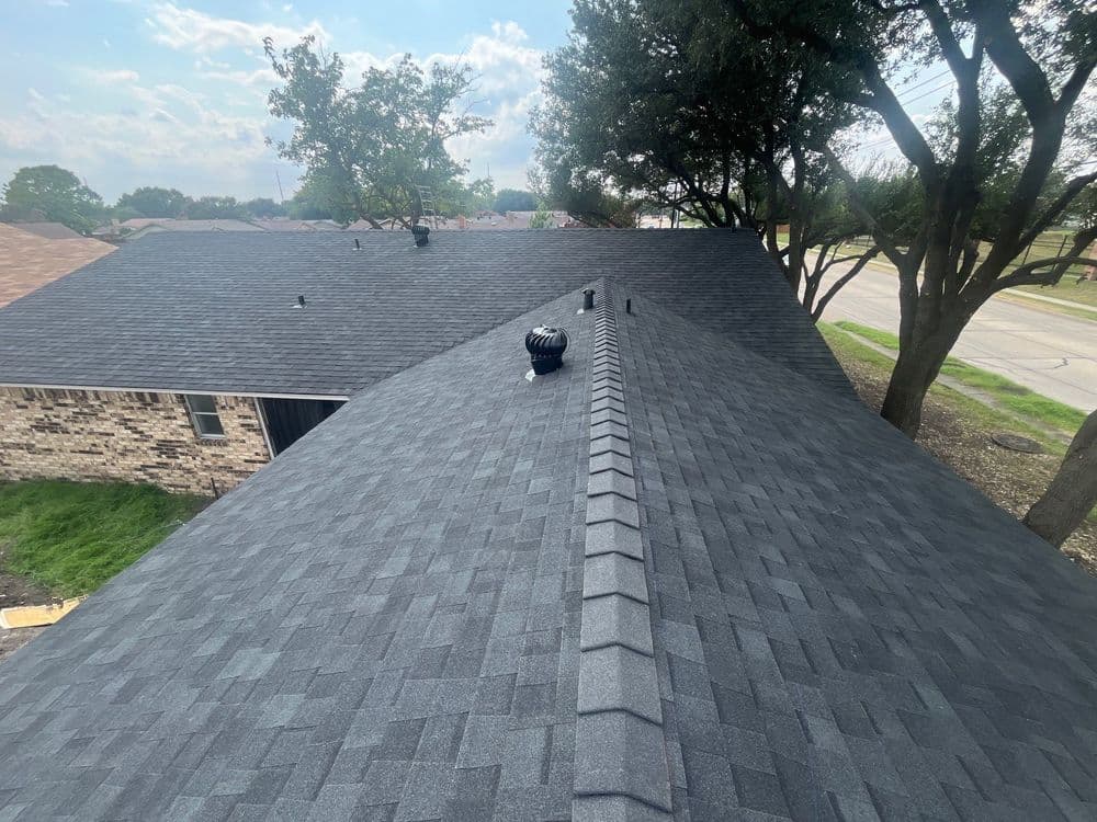 Aerial view of a dark shingle roof with vents and surrounding trees on a sunny day.
