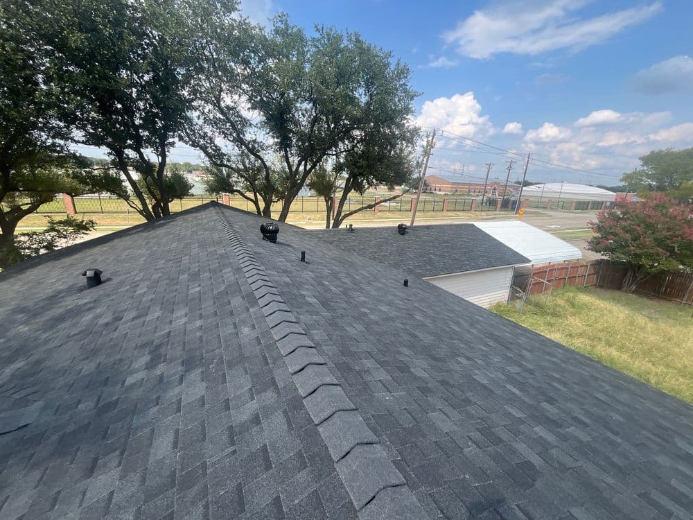 Aerial view of a dark shingle roof with trees and blue sky in the background.