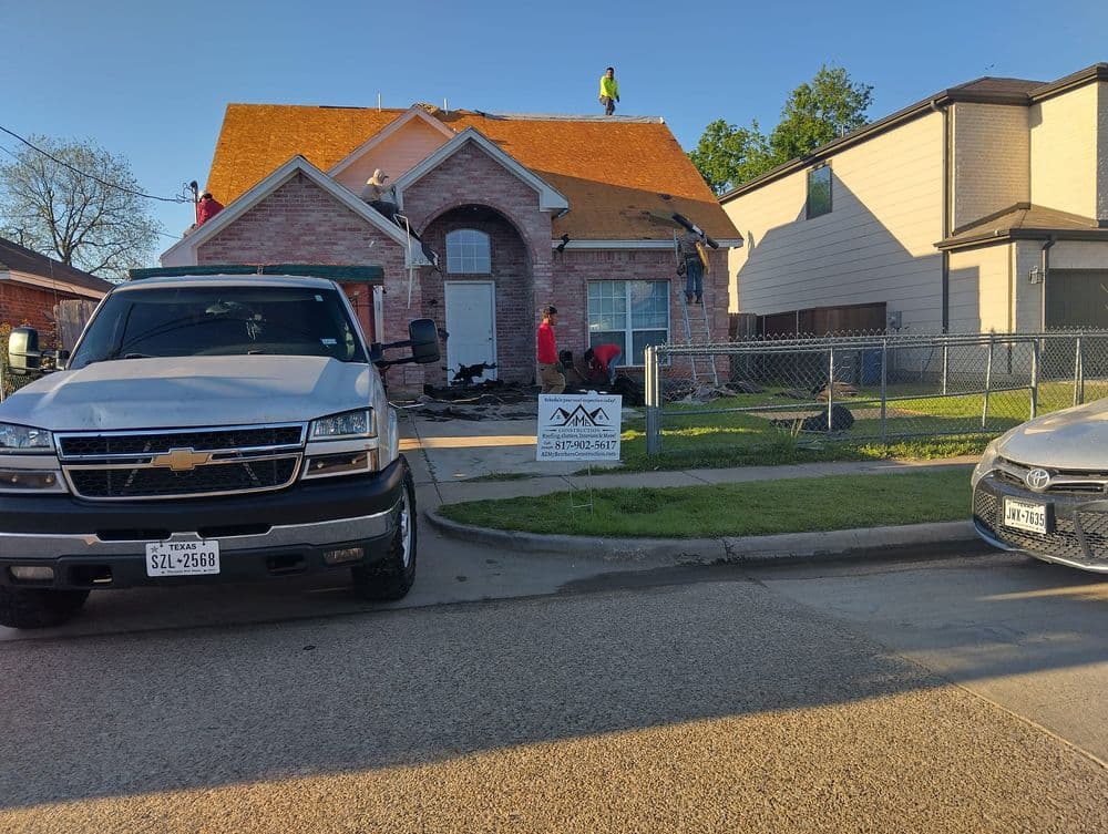 Roofing crew working on a house with orange shingles and parked trucks nearby.