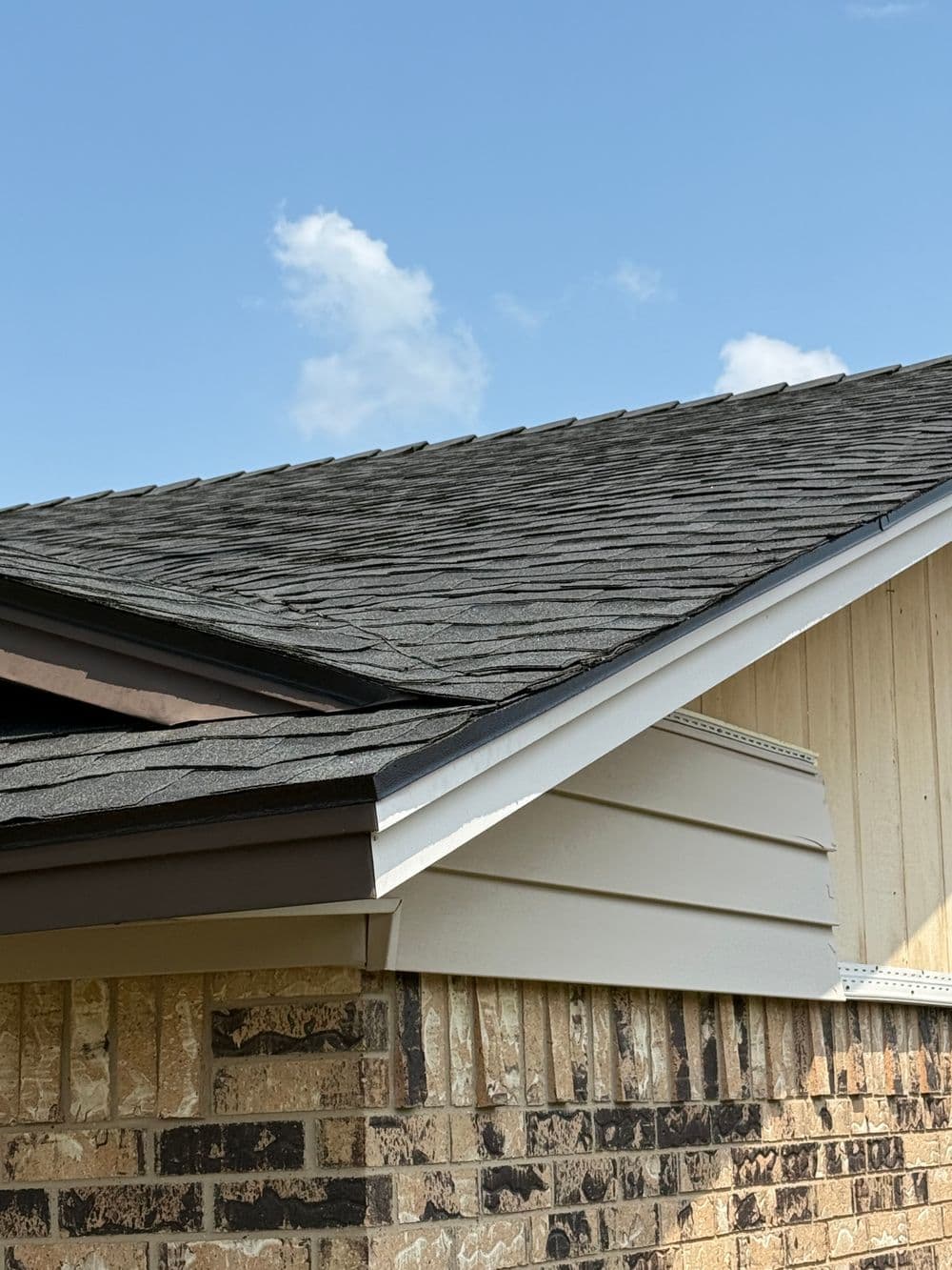Detailed view of a residential roof with shingles, eaves, and brick siding against a blue sky.