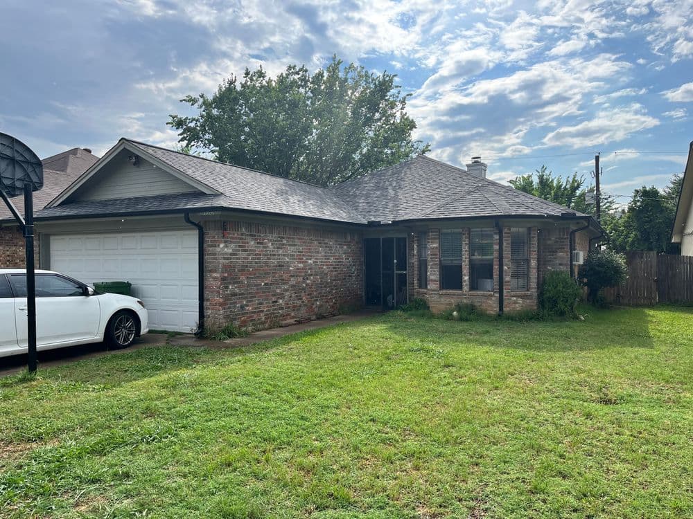 Brick house with a front lawn, driveway, and basketball hoop under a partly cloudy sky.